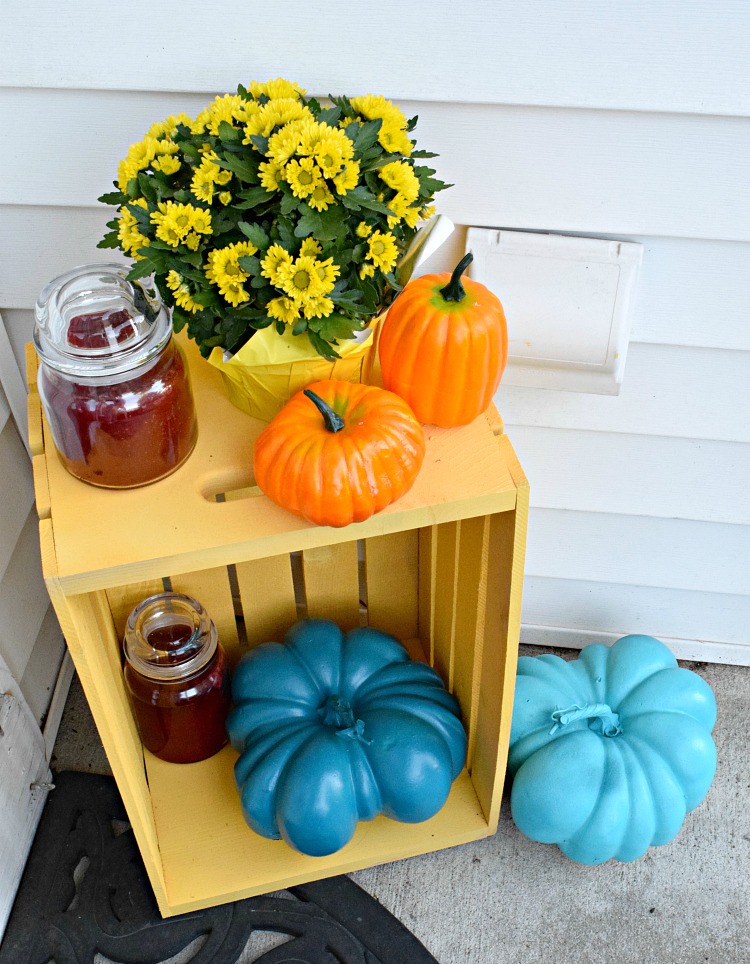 A front porch fall display that includes two blue pumpkins, mini orange pumpkins, a yellow crate, mums and candles.