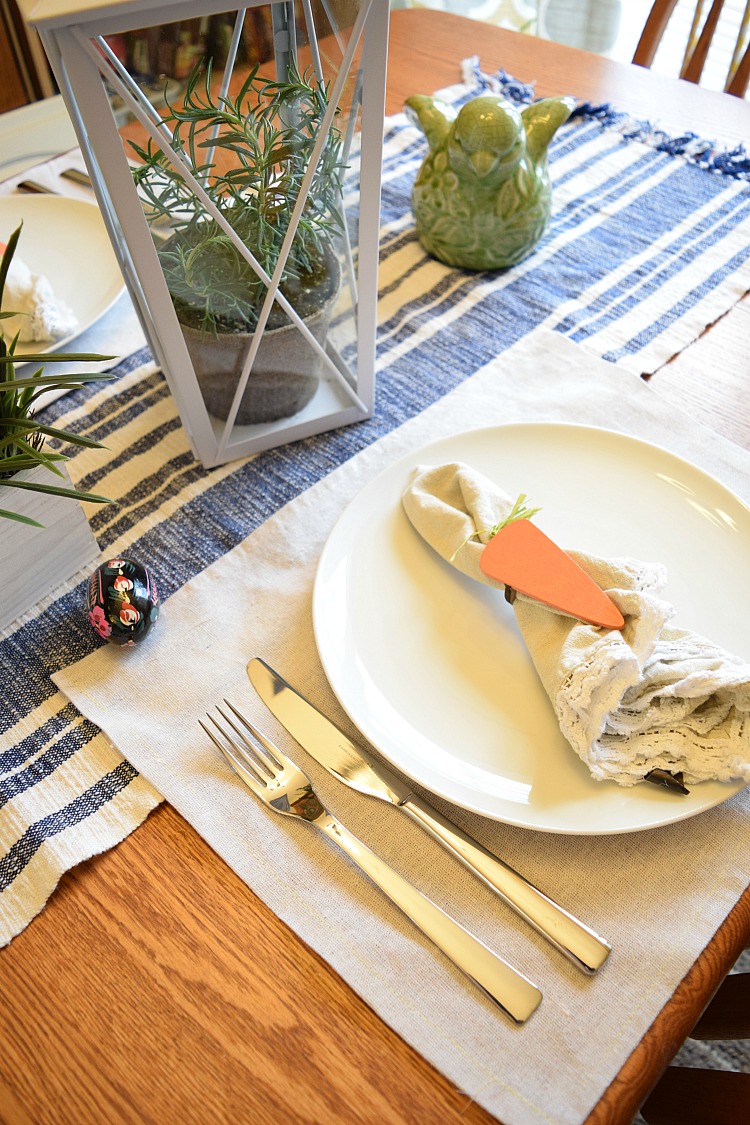 An Easter tablescape with a blue and white striped table runner, white porcelain dishware and stainless steel, and a white lantern with potted rosemary