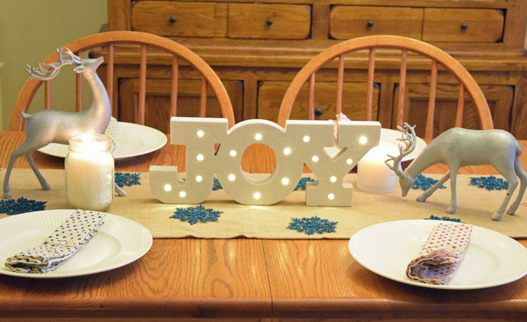 A Christmas table with silver reindeer, a burlap table runner, white lanterns and a lighted marquee joy sign