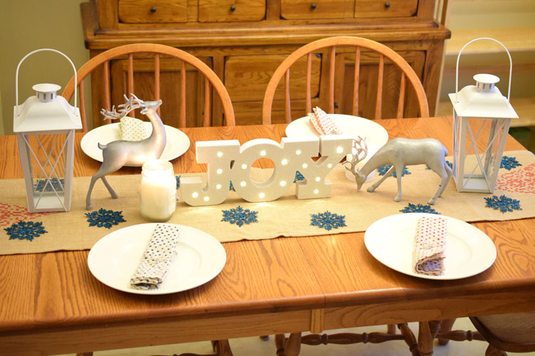 Christmas farmhouse table with burlap table runner, silver reindeer, candles and lanterns, plus a lighted Joy marquee sign