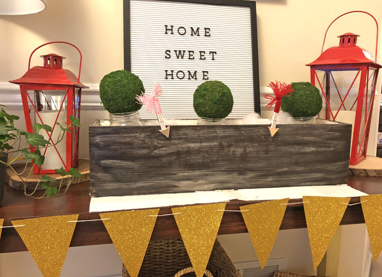 Console table decorated for Valentine's Day with a gold banner, red lanterns, planter box, bell jars filled with moss and a letter board.