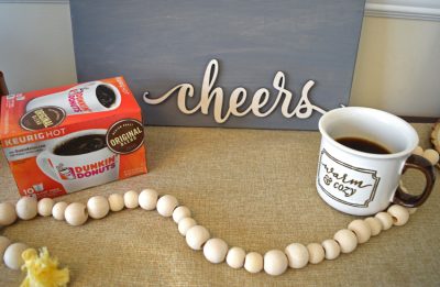 cup of coffee on a coffee table in front of a cheers sign and behind a wooden bead garland