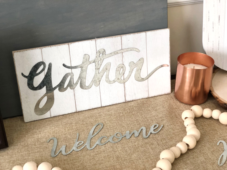 metal welcome word, gather farmhouse-style sign, copper candle and natural wood bead garland on a fall console table