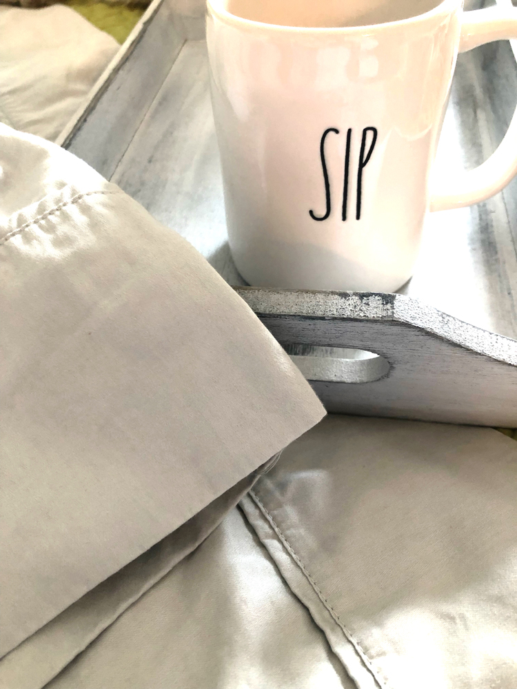 Gray breakfast tray with floral eyeglass case, Rae Dunn sip mug and mini blue teapot on a set of cotton gray sheets