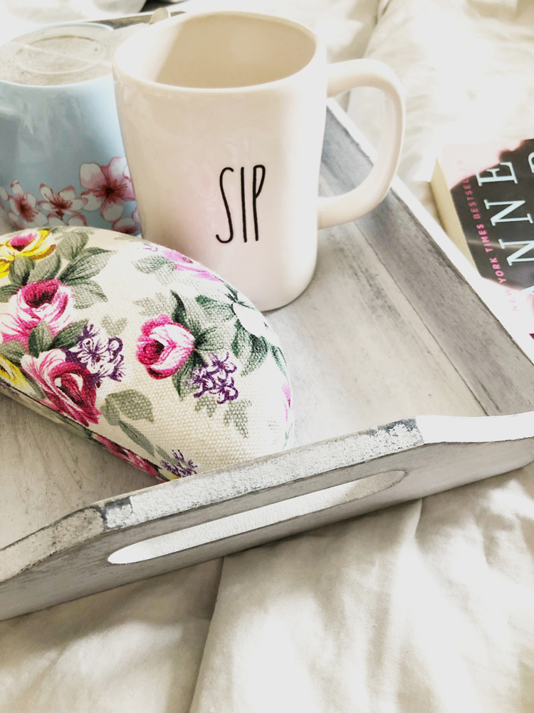 Gray breakfast tray with floral eyeglass case, Rae Dunn sip mug and mini blue teapot on a set of cotton gray sheets