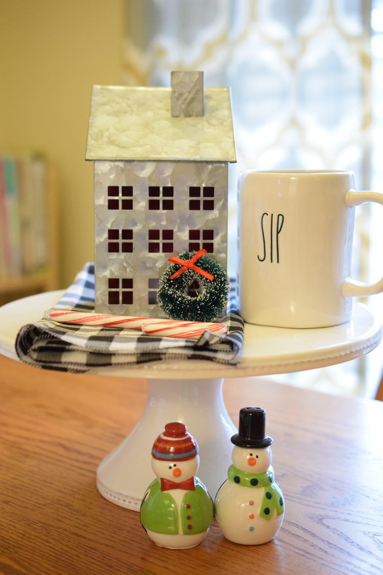 cake stand with a Rae Dunn mug, galvanized tin house, buffalo plaid napkin, candy cane, mini wreath and snowmen salt and pepper shakers.