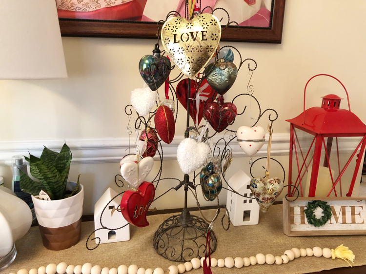 A Valentine's Day console table with a heart ornament tree filled with heart ornaments. Also on the table are a wooden bead garland, a potted snake plant , a home sign and a red lantern.