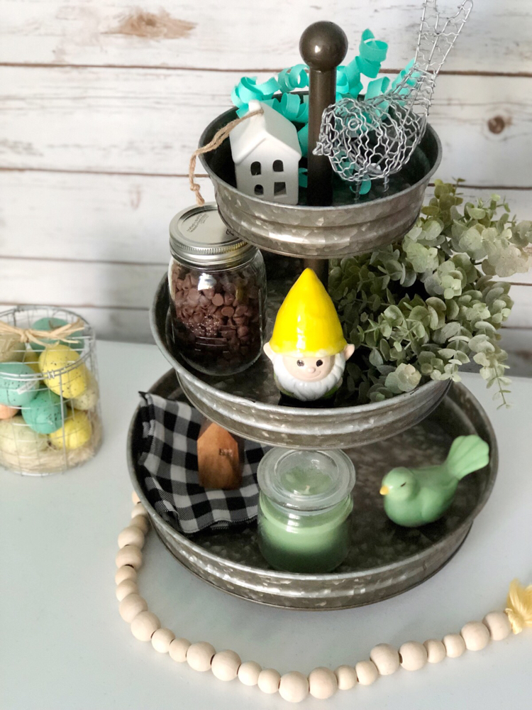 A galvanized tiered tray for spring decorated with birds, a garden gnome, miniature houses and faux greenery.