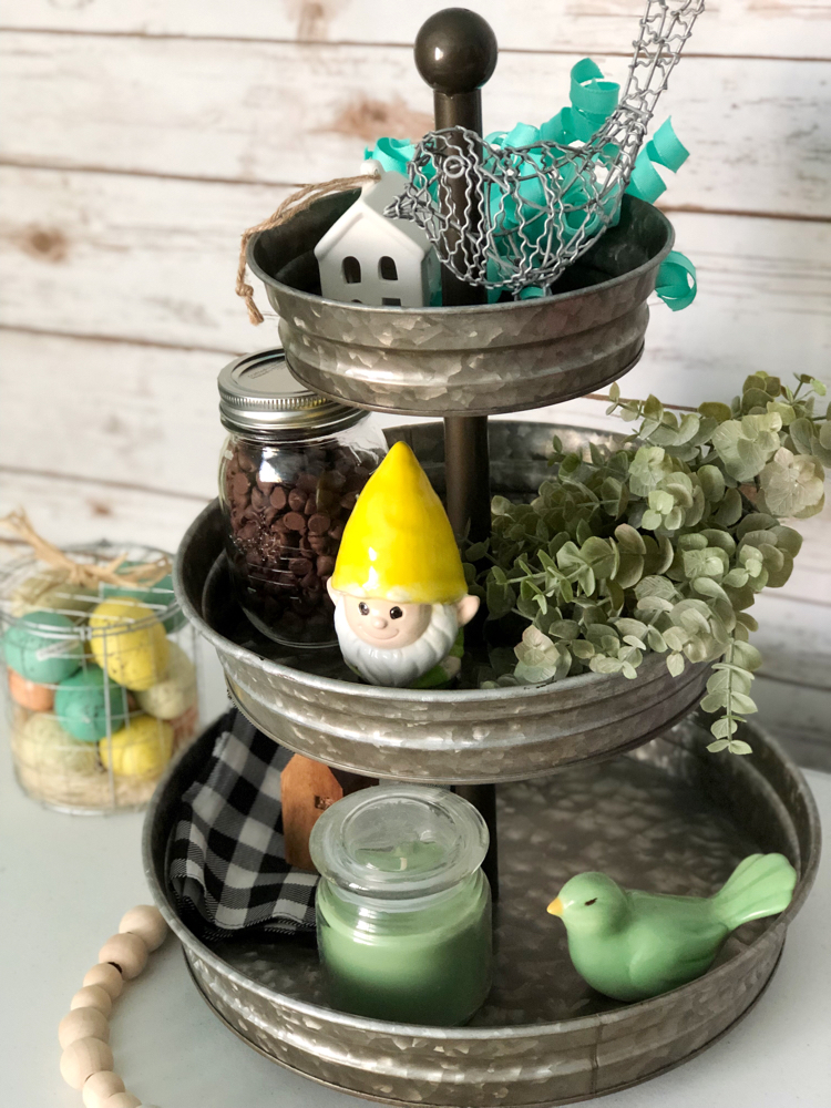 A three tiered galvanized tray decorated for spring with faux greenery, a garden gnome, ceramic and wire birds, mini houses, a buffalo plaid napkin and a candle.