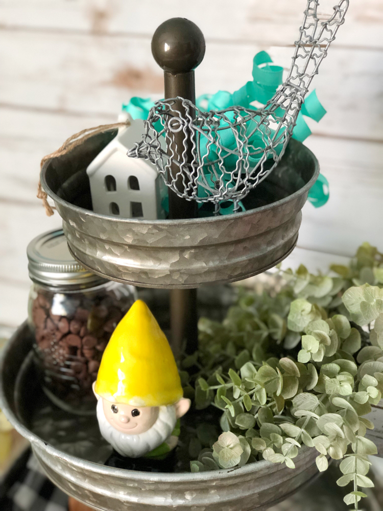 A galvanized metal tiered tray with miniature birds and houses, a garden gnome, faux greenery and a buffalo plaid napkin.