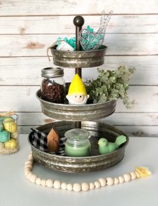 A three tiered galvanized decorated for spring with faux greenery, a garden gnome, ceramic and wire birds, mini houses, a buffalo plaid napkin and a candle.