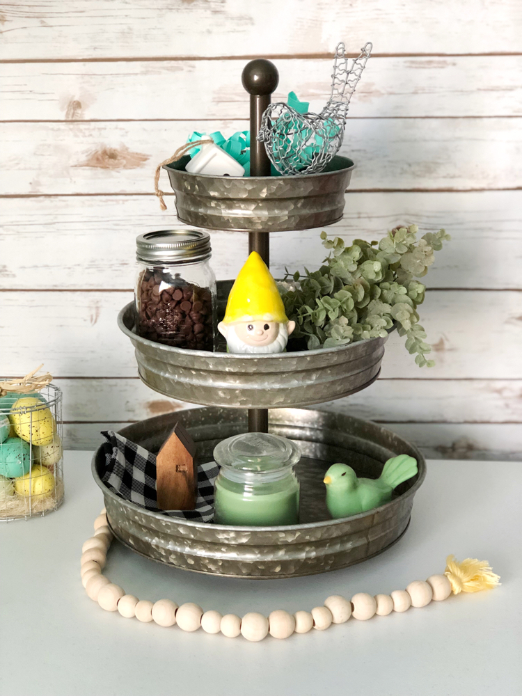 A three tiered galvanized decorated for spring with faux greenery, a garden gnome, ceramic and wire birds, mini houses, a buffalo plaid napkin and a candle.