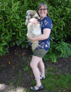 Woman in a blue floral top wearing olive shorts and grey sandals and holding a Shih Tzu.