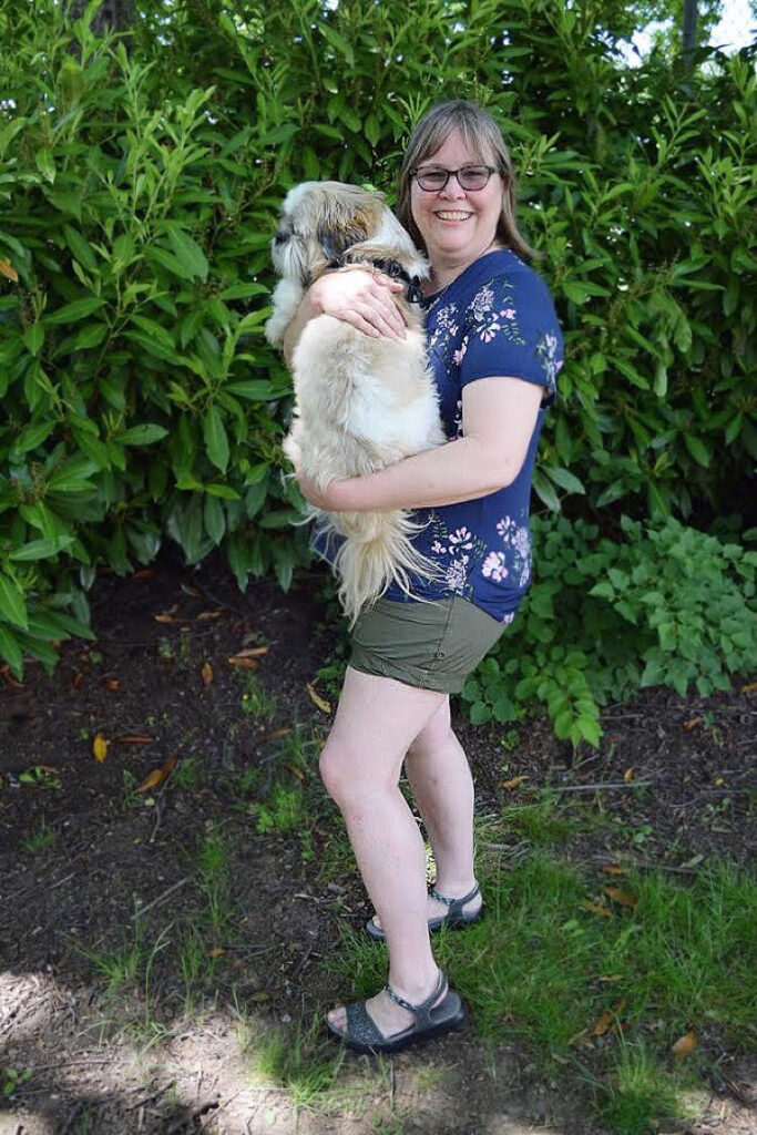 Woman in a blue floral top wearing olive shorts and grey sandals and holding a Shih Tzu.