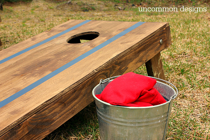 Cornhole board stained brown with painted blue stripes. A bucket of beanbags is next to the board. Photo by Uncommon Designs