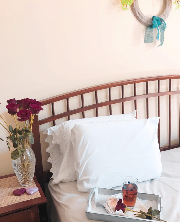 A farmhouse style spindle bed with white sheets. A pretty burlap and floral wreath is on the wall and vase of roses is on the nightstand.