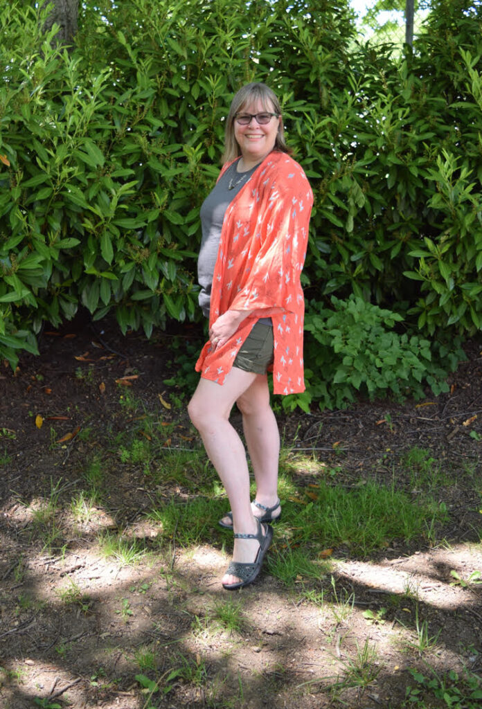 A woman models a peach colored kimono, a gray T shirt, olive shorts and gray sandals.