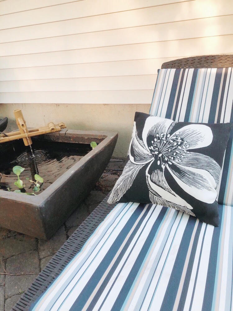 Chaise lounge with a blue and white striped cushion and a black pillow with a flower print. A patio pond with a fountain and water hyacinths is nearby on the paver patio.