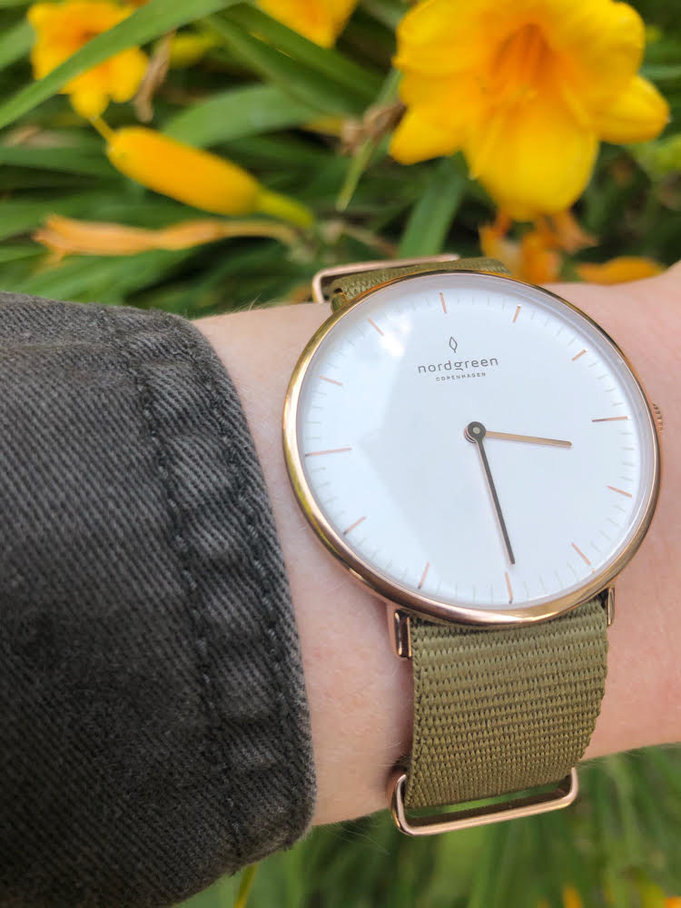 A Nordgreen rose gold watch with a green nylon strap against some pretty yellow lilies