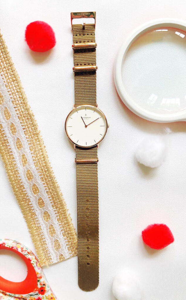 Watch with craft supplies surrounding it - a magnifying class, pom pots and burlap ribbon accented with lace