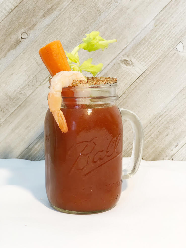 A Bloody Mary in a Ball mason jar glass. The drink is garnished with celery, a carrot stick and a shrimp.