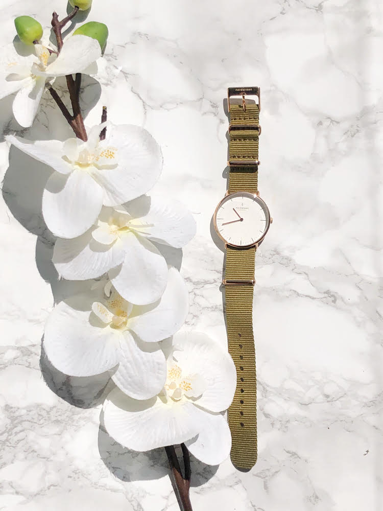 A rose gold watch with a white face and a green nylon band on a marble surface with white flowers