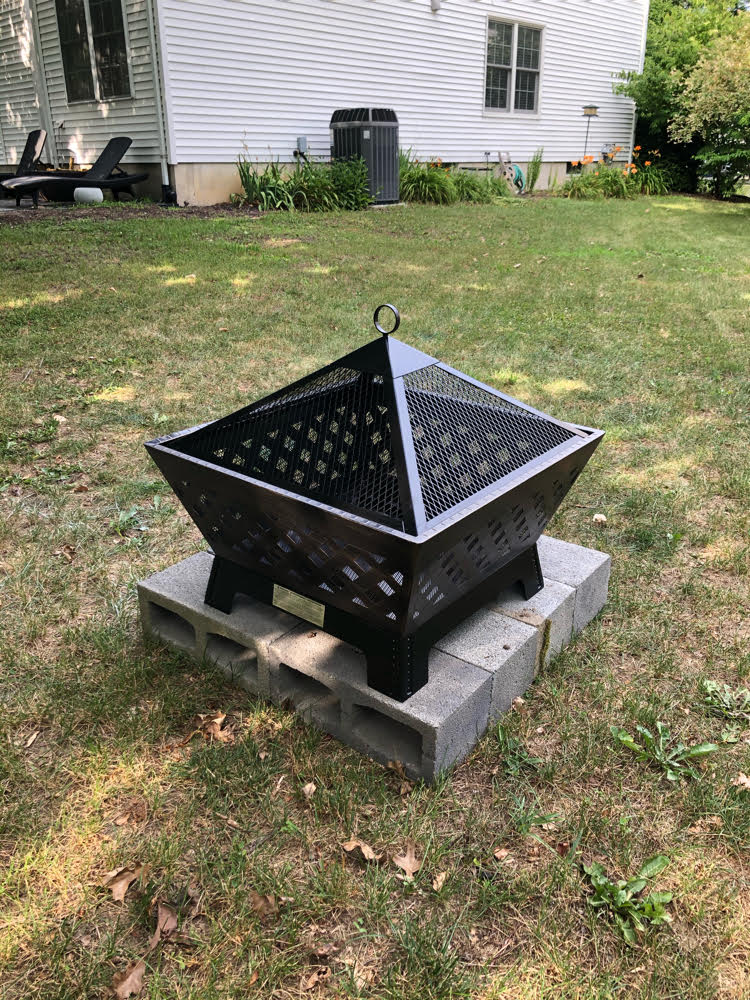 An antique bronze Landmann 24" fire pit set on a square of concrete blocks in a backyard.