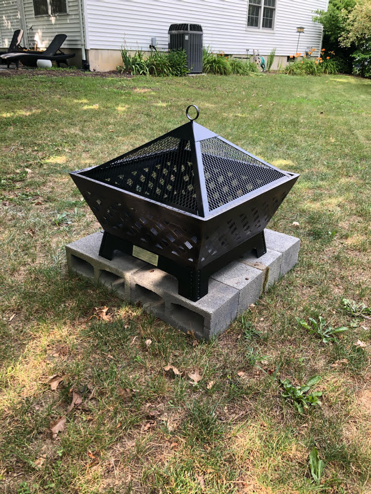 A Landmann metal fire pit with a spark screen sitting on a DIY base of concrete blocks in a backyard