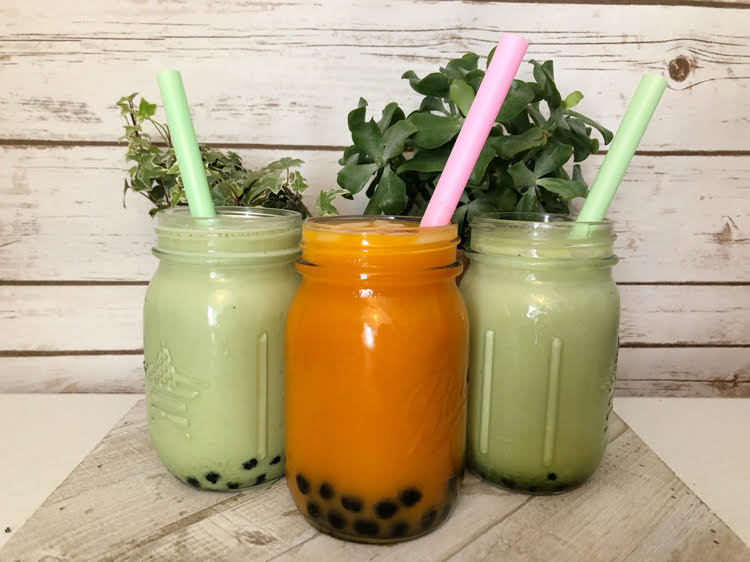 a mason jar of homemade Thai bubble tea and two mason jars of matcha green tea against potted ivy and a planked farmhouse wall