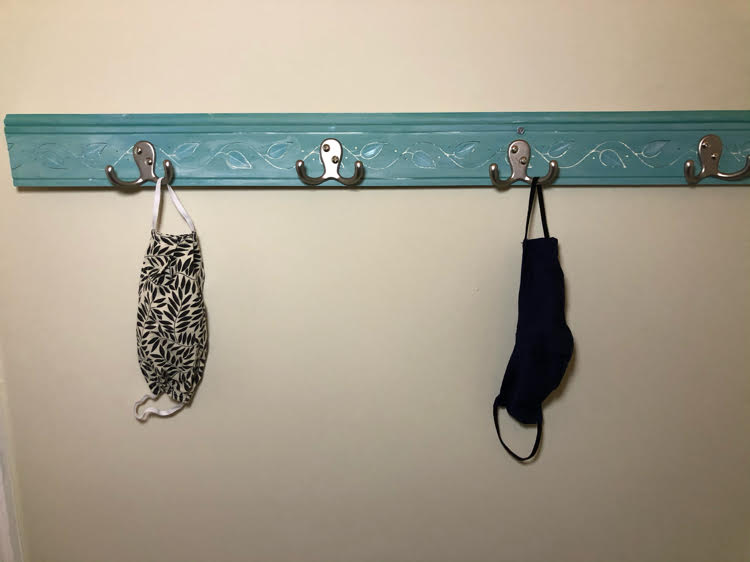 A DIY face mask rack made with double coat hooks, pretty chair rail molding, and spray painted a beautiful blue green