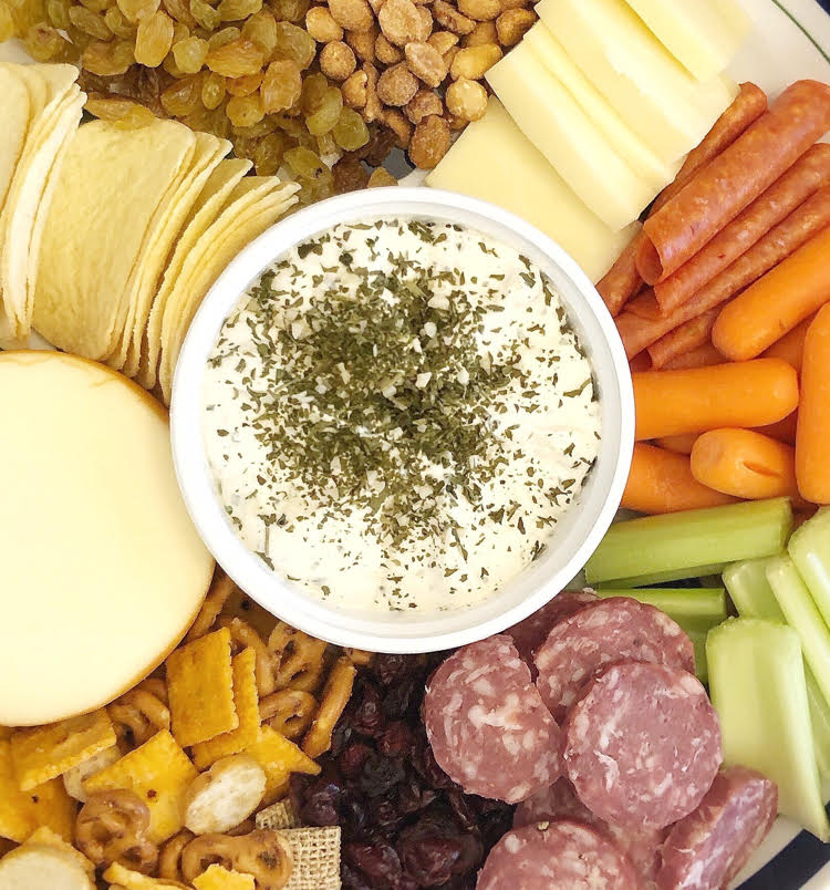 garlic and herb soft Alouette Cheese on a charcuterie board with meats, celery and carrot sticks, peanuts, raisins and potato chips