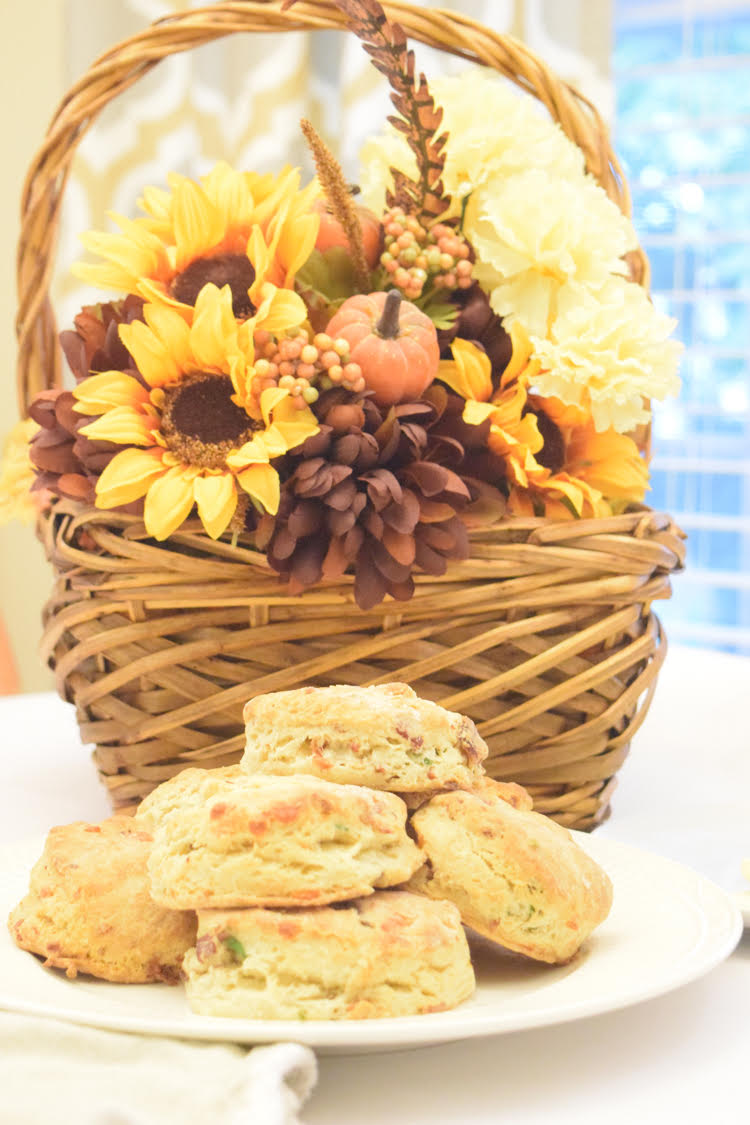 bacon cheddar chive biscuits in front of a plate, sitting in front of a wicker basket holding a faux floral and fall leaf arrangement