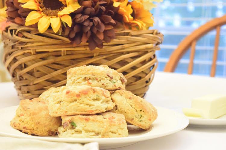 delicious bacon cheddar chive biscuits on a white plate, in front of a wicker basket with a fall arrangement of flowers and fall leaves