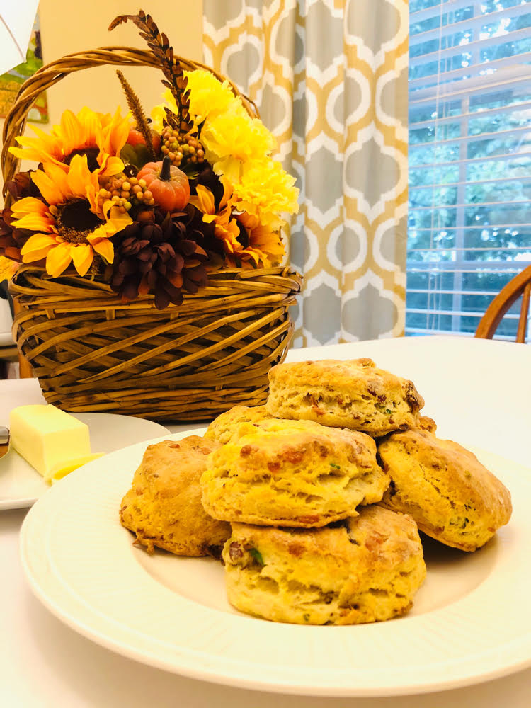 cheddar biscuits with cheddar cheese and chives stacked on a plate in front of a basket with an arrangement of faux fall flowers