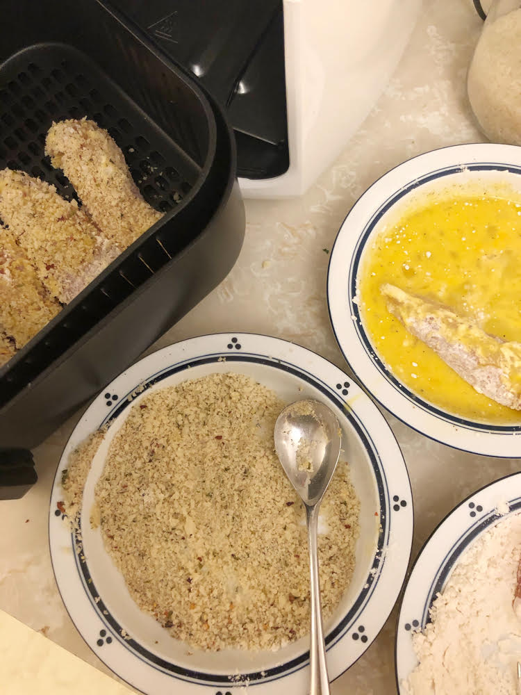 breaded chicken in the basket of an air fryer with bowls of breadcrumbs, egg and flour next to the air fryer