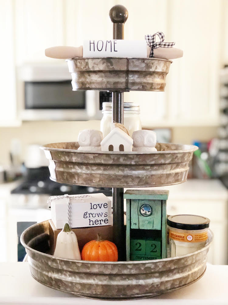 a galvanized 3-tier tray in a kitchen. On the tray on owl salt and pepper shakers, pumpkin and squash salt and pepper shakers, a wooden birdhouse calendar, a jarred candles, a ceramic house a mini "home" rolling pin and a "love grows here" faux book stack.