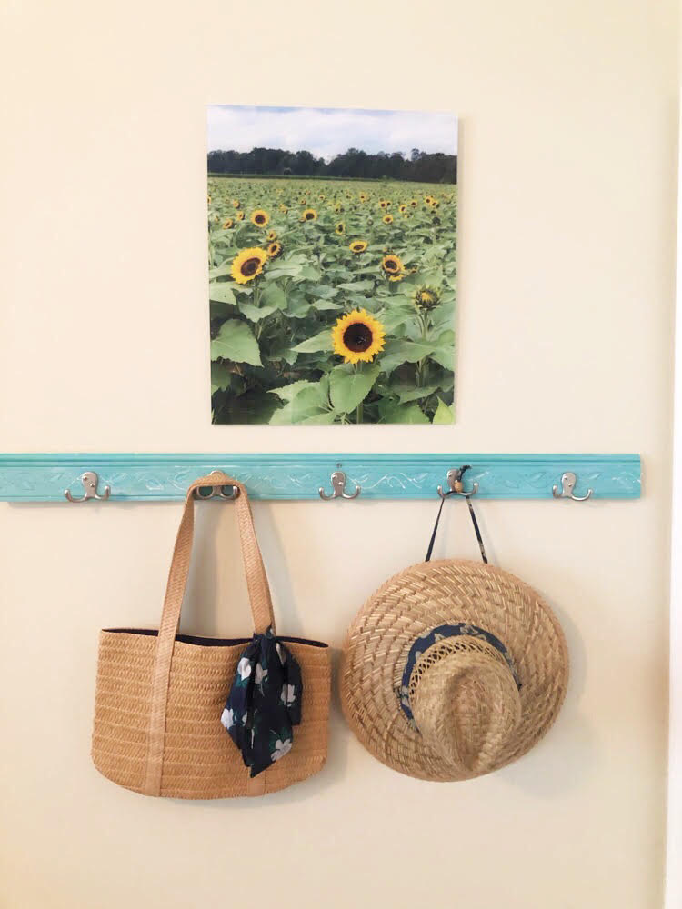 A straw tote and a straw hat hanging on a DIY coat rack under a pretty print of a sunflower field.