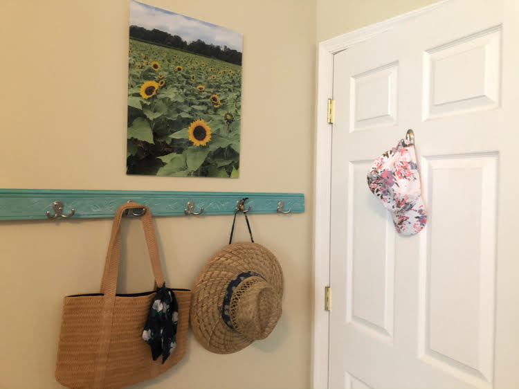 acrylic print of a sunflower field hanging over a coat rack holding a straw bag and hat by a white door