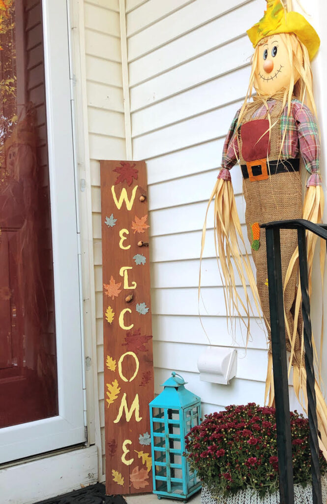a small front porch decorated for fall with a DIY standing welcome sign, mums, a blue lantern and a scarecrow