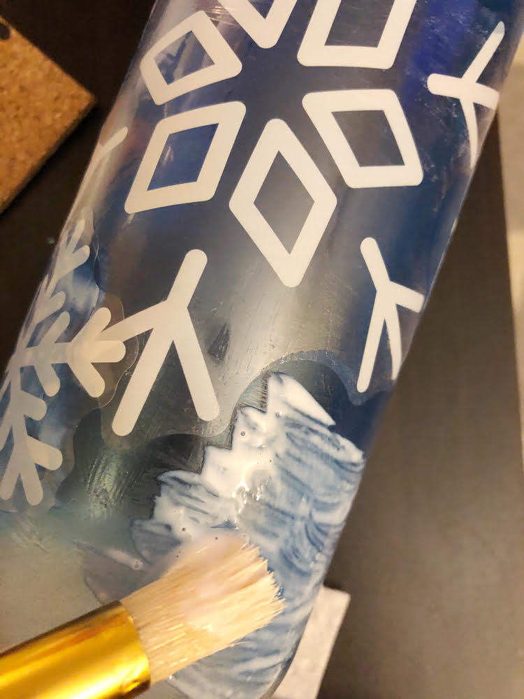 gluing white vinyl snowflake clings to a blue wine bottle with Mod Podge and a stencil brush