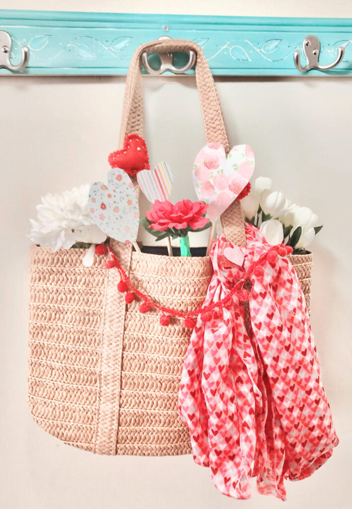 A pretty straw tote decorated for Valentine's Day with a pom pom garland, heart scarf, faux flowers and felt and paper hearts.