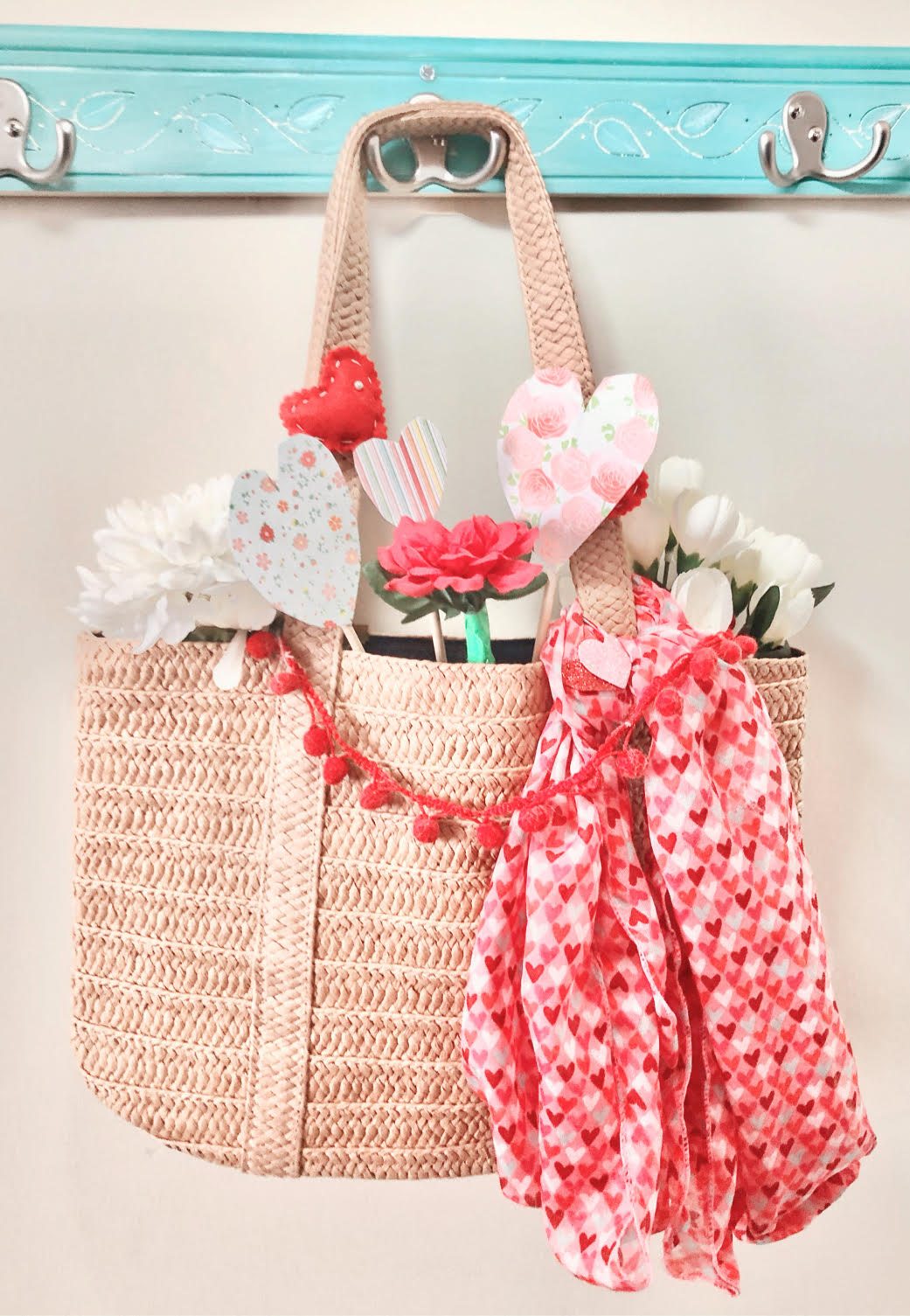 A pretty straw tote decorated for Valentine's Day with a pom pom garland, heart scarf, faux flowers and felt and paper hearts.