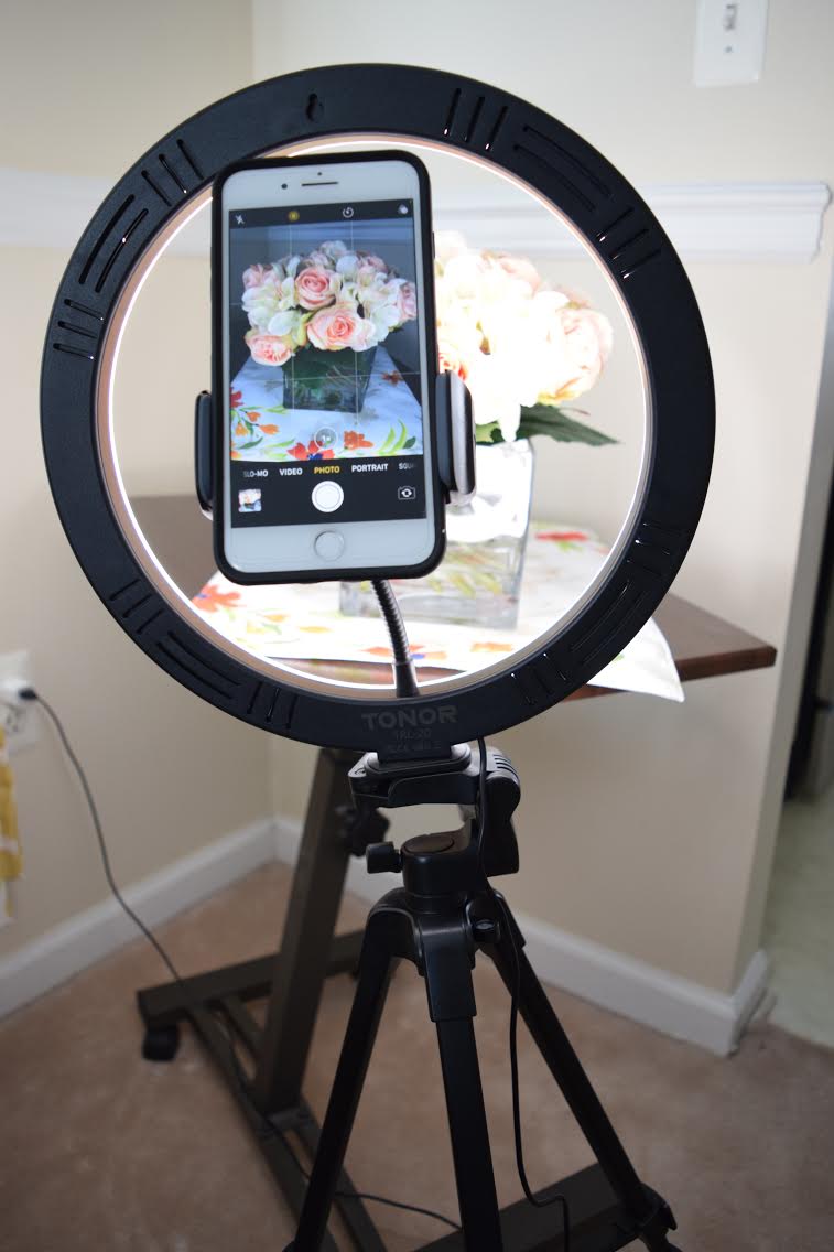 A mini photo studio setup with an iphone, a TONOR 12 inch selfied ring light with tripod stand, smartphone holder and Bluetooth Remote Shutter, and a Seville mobile laptop desk cart