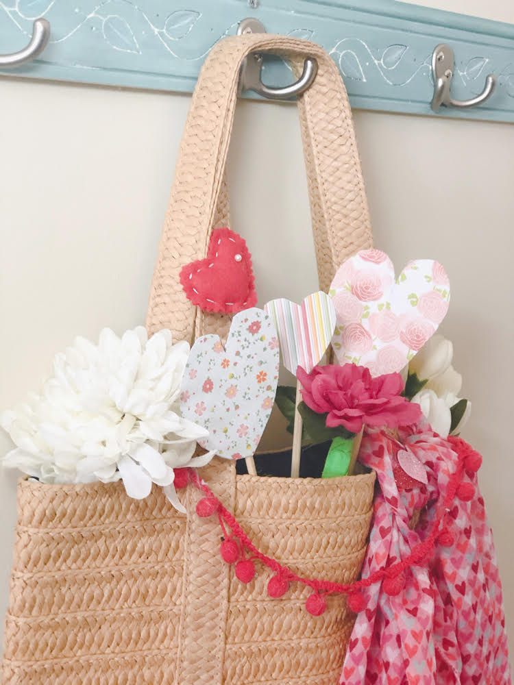 A straw tote decorate with faux flowers, felt and paper hearts, a heart scarf and a pom pom garland.