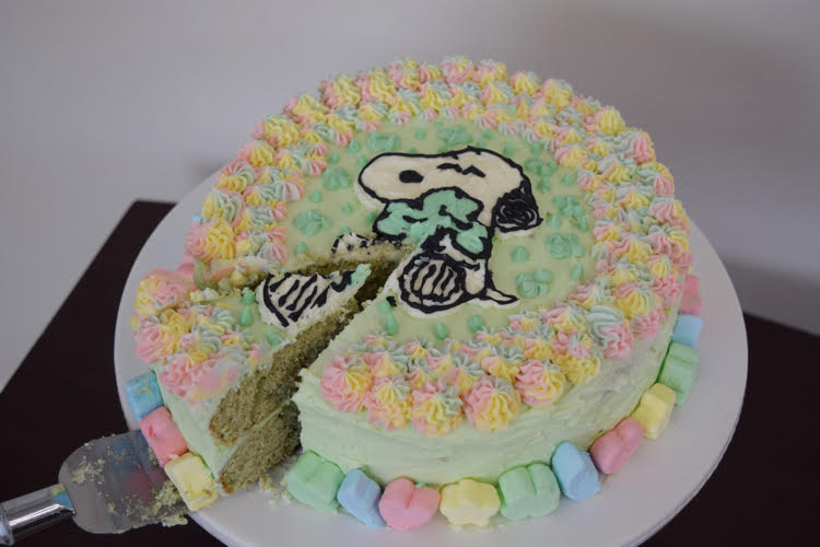 A green frosted cake with rainbow piped stars and a Snoopy and shamrock design on the top. The cake is lined with Lucky Charms marshmallows