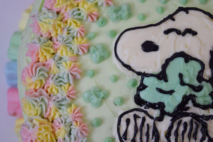 A matcha layer cake decorated with a Snoopy holding a shamrock design and rainbow icing.